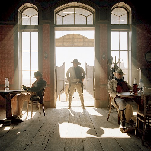 A cowboy stands in a saloon doorway, silhouetted by sunlight, with two men seated at tables on either side, creating a classic Western scene.