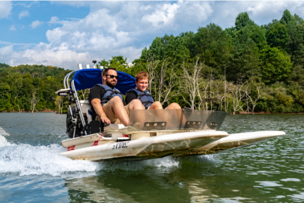 Two people are riding a motorized watercraft on a lake, wearing life vests. The backdrop is a lush, green forest under a cloudy sky.