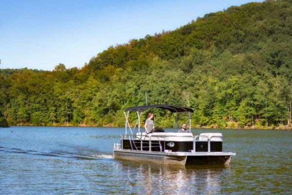 A pontoon boat with people on board glides across a calm lake surrounded by lush green trees under a clear blue sky, creating a serene scene.