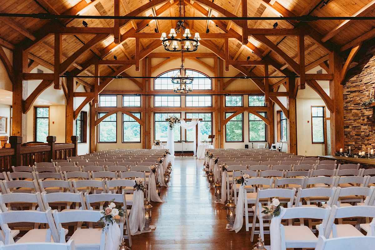 This is a decorated wedding venue with rows of white chairs facing an altar, set in a rustic wooden building with large windows and chandeliers.