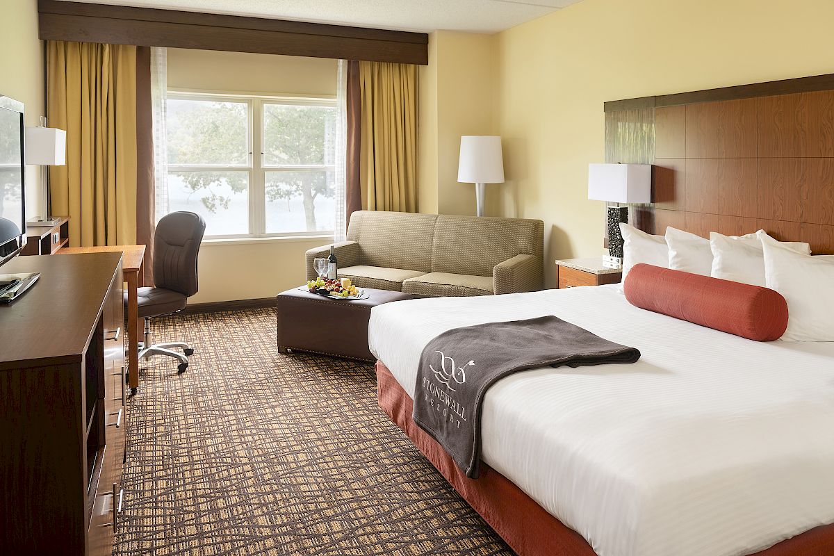 This image shows a well-lit hotel room with a bed, sofa, work desk, and TV, featuring large windows and warm-toned decor.
