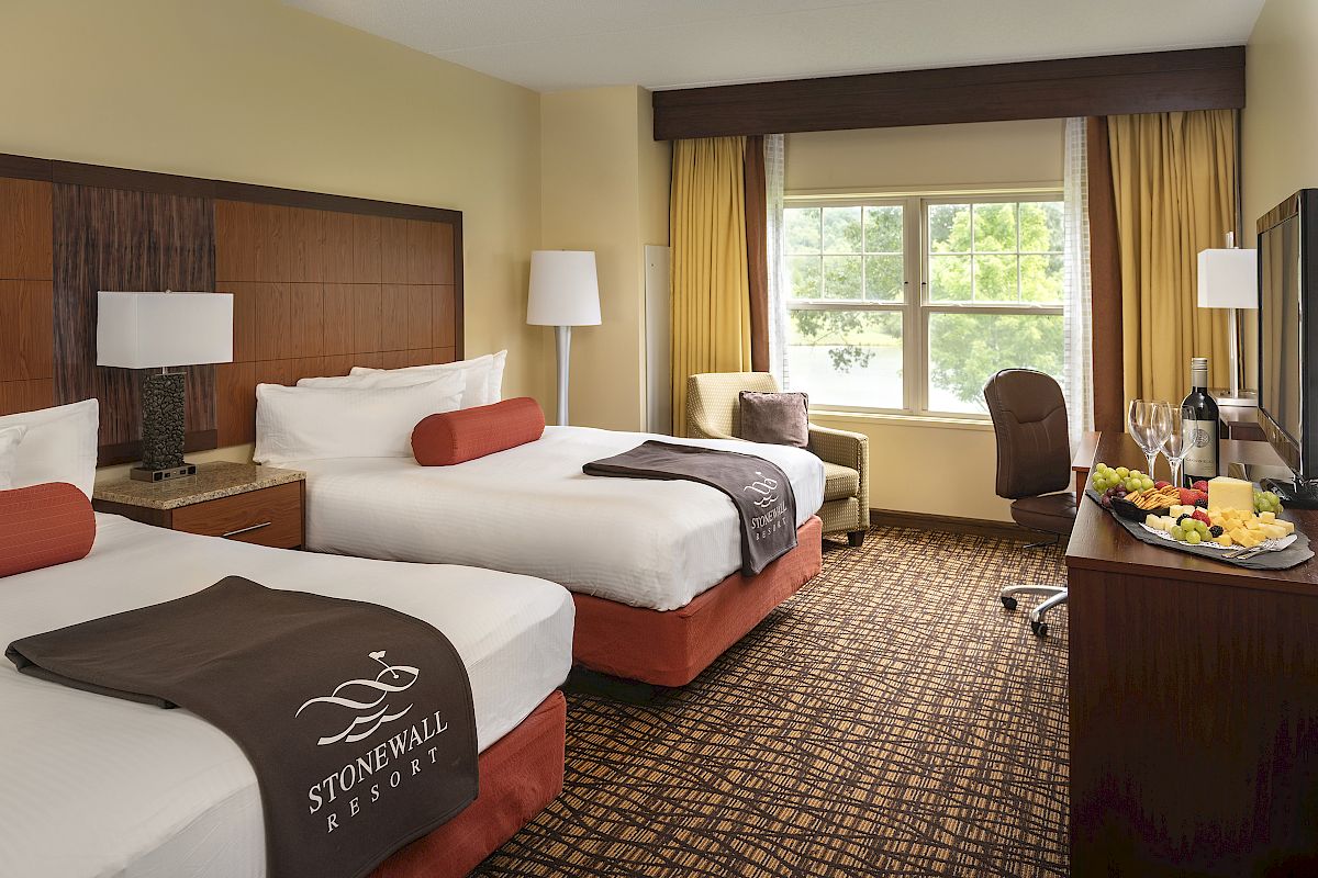 A hotel room with two beds, window, chairs, table, lamp, and a TV. Beds have linens labeled