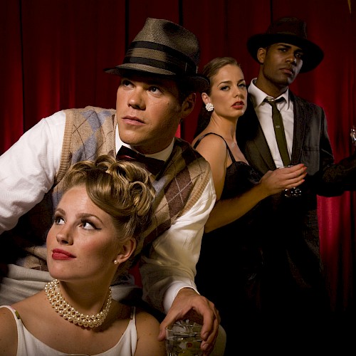 Four people dressed in vintage attire with hats and dresses pose against a red curtain backdrop, holding drinks.