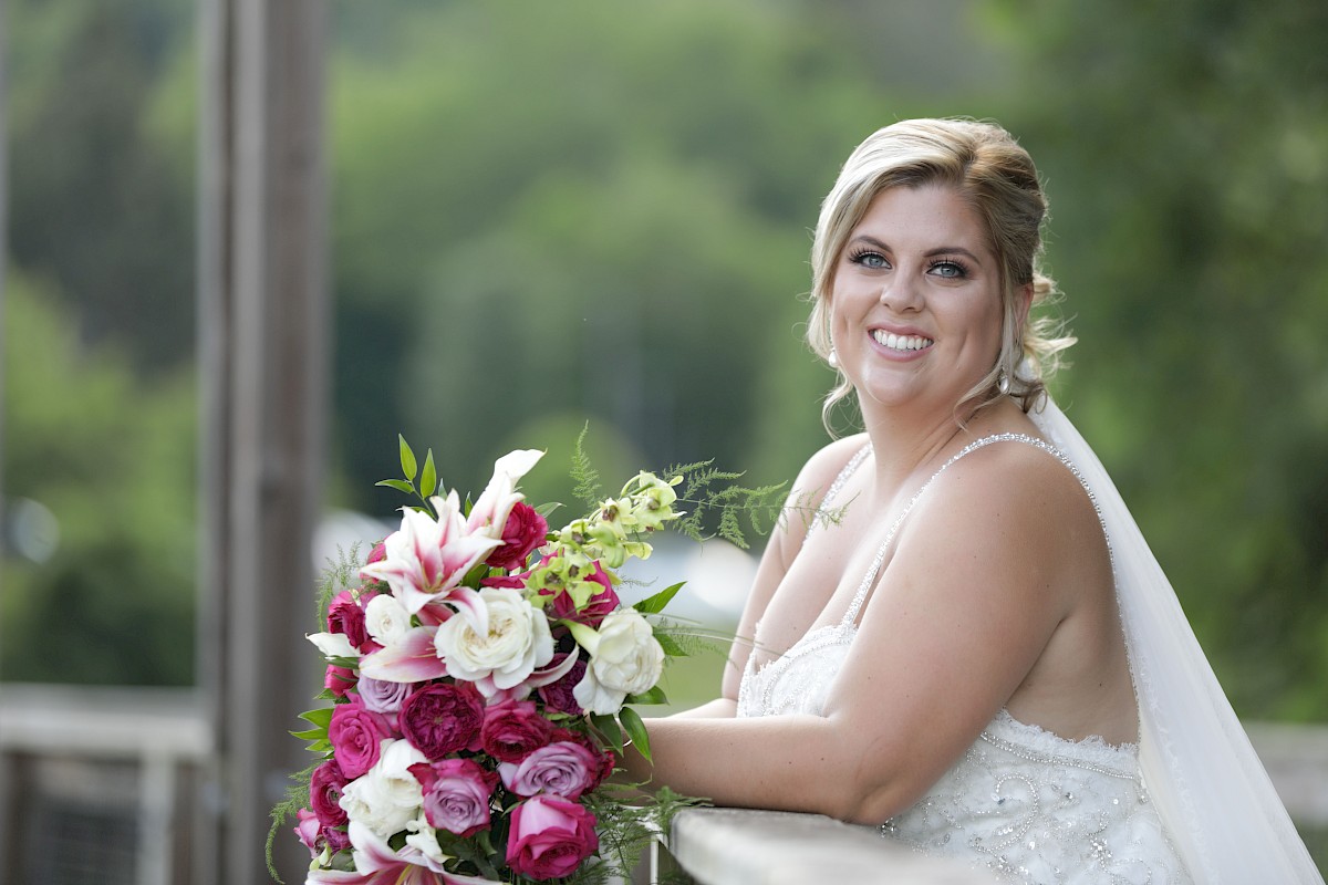 A bride holding a bouquet of pink and white flowers stands on a bridge, smiling with a blurred green background.