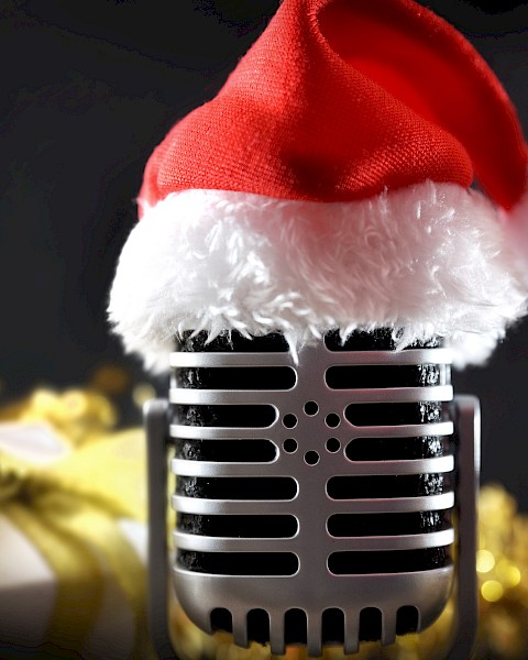 A vintage microphone wearing a Santa hat is in focus, with festive decorations and a gift in the blurred background.