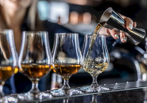 A person is pouring drinks into a row of glasses using a jigger, likely preparing cocktails or shots at a bar.