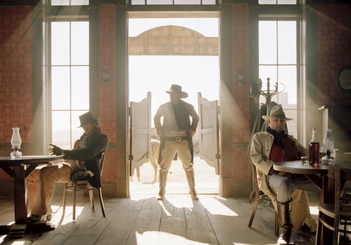 A cowboy stands in a saloon doorway, silhouetted by sunlight, with two men seated at tables on either side, creating a classic Western scene.