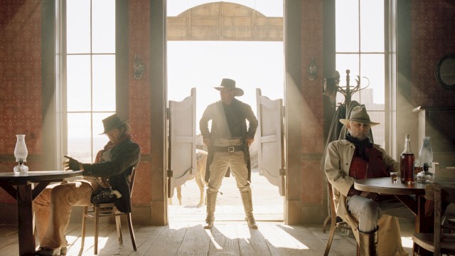 A cowboy stands in a saloon doorway, silhouetted by sunlight, with two men seated at tables on either side, creating a classic Western scene.
