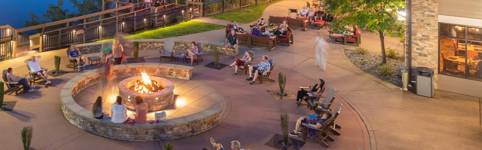 A scenic outdoor area with people sitting around a fire pit near a lake, surrounded by trees and string lights, next to a building with lights.