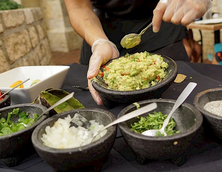 A person is making guacamole with bowls of ingredients, including onion, cilantro, green onions, tomatoes, and spices, on a table.