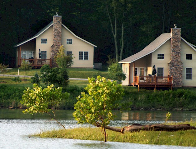 The image shows two houses with stone chimneys near a lake, surrounded by trees and greenery.