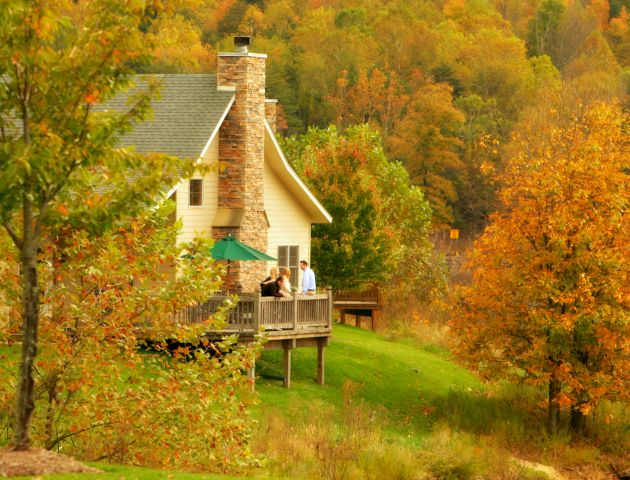 A cozy cabin with a stone chimney is surrounded by autumn foliage. People are standing on the deck enjoying the view in the peaceful, scenic setting.