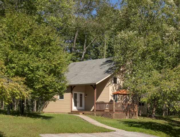 A small house with a gray roof and beige siding, surrounded by lush green trees, with a pathway leading to the front door ending the sentence.