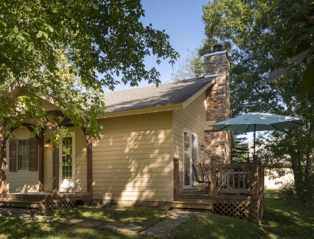 A cozy yellow house with a stone chimney, surrounded by trees. There's a small deck with an umbrella. The scene is peaceful under a clear sky.