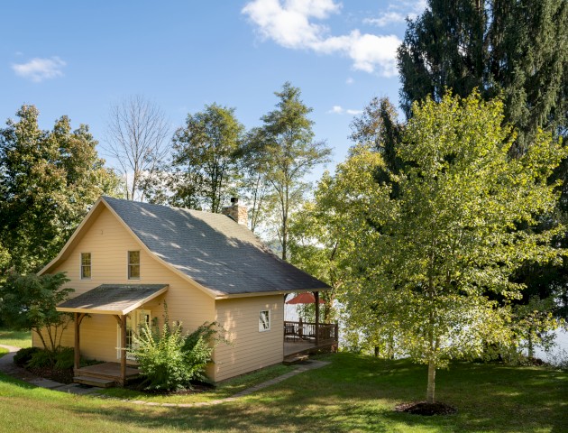 The image shows a charming small house surrounded by lush greenery and trees, with a clear blue sky in the background, indicating a pleasant day.