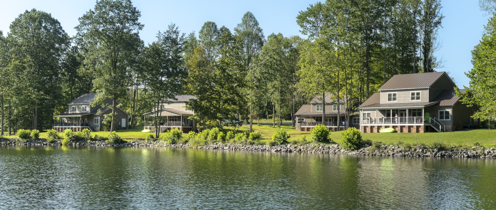 Several houses are situated along a tranquil lakeside, surrounded by lush trees and greenery, reflecting a peaceful and serene environment.