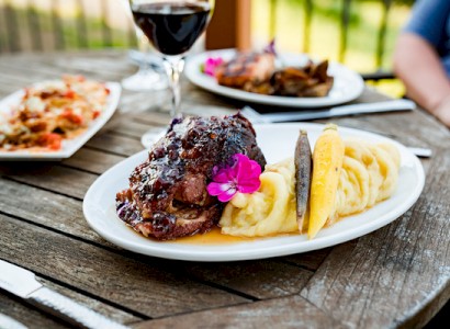 A plate with roasted meat, mashed potatoes, and a garnish, along with a glass of red wine on a rustic outdoor table.