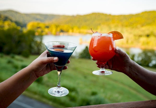 Two people are holding colorful cocktails outdoors with a scenic river and green hills in the background.