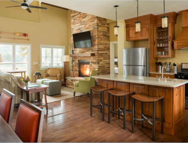 A cozy open-concept living room and kitchen with wooden cabinets, a stone fireplace, and modern appliances. The room has warm, inviting decor.