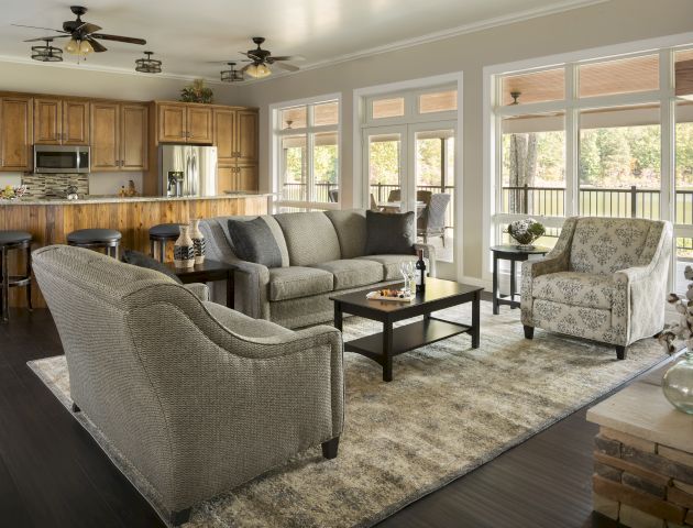 A modern open-concept living room and kitchen with grey seating, wooden cabinets, bar stools, a rug, and large windows facing outdoors end the sentence.