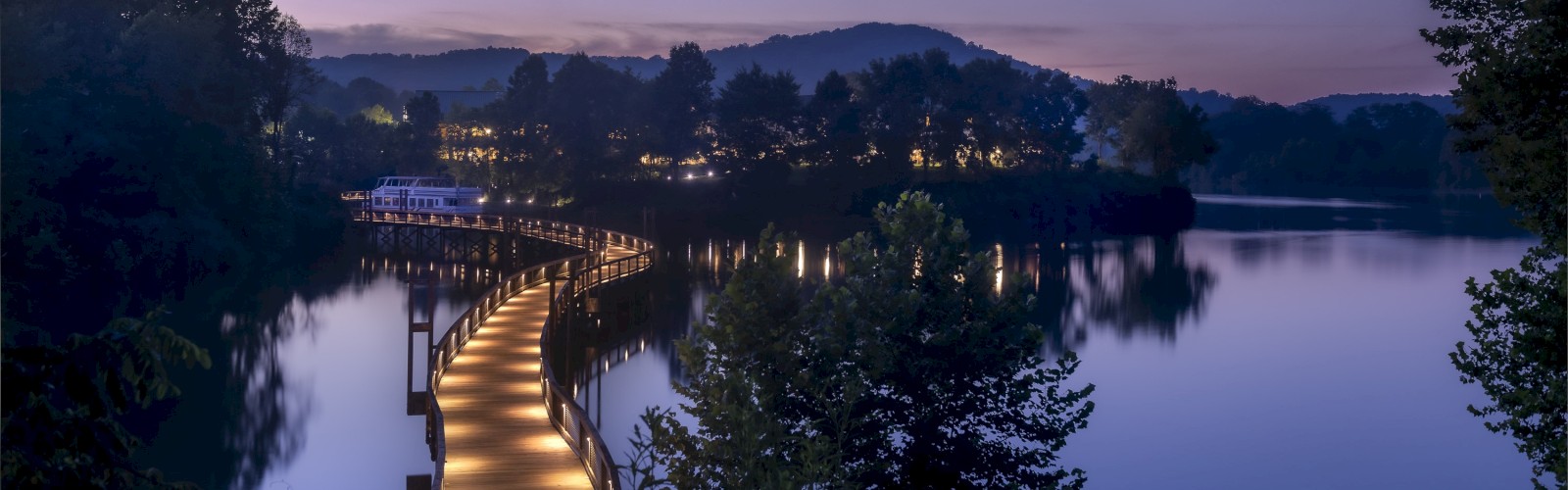 A wooden walkway over calm water, surrounded by trees and hills during twilight, with soft lights illuminating the path, ending the sentence.