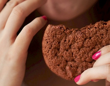 A person is biting into a crumbly chocolate cookie with pink nail polish on fingernails, close-up shot.