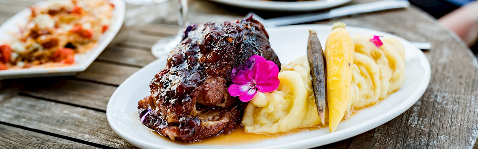A plate with mashed potatoes, meat topped with glaze, garnished with edible flowers, and a glass of red wine on a wooden table.