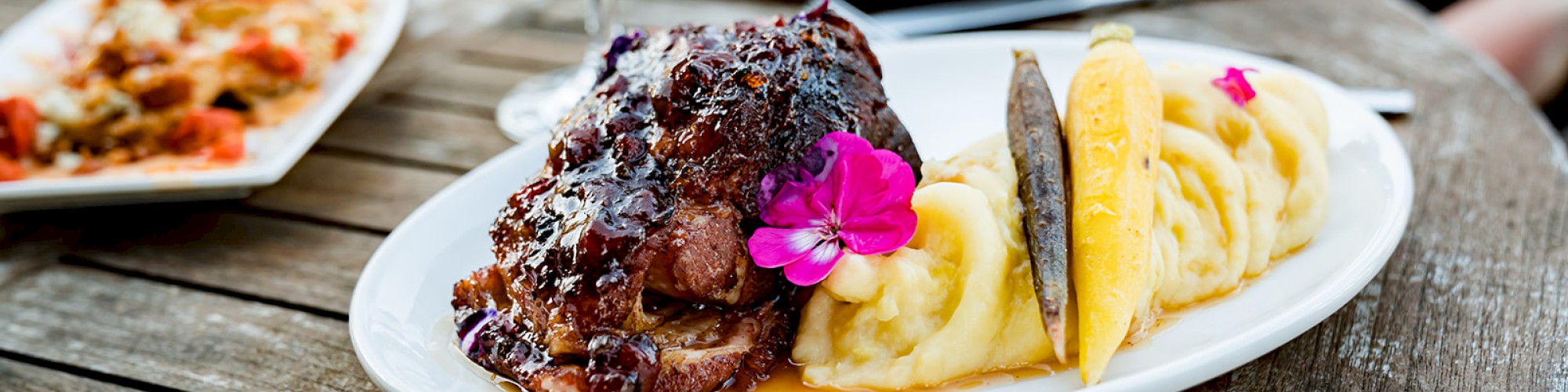 A plate with mashed potatoes, meat topped with glaze, garnished with edible flowers, and a glass of red wine on a wooden table.