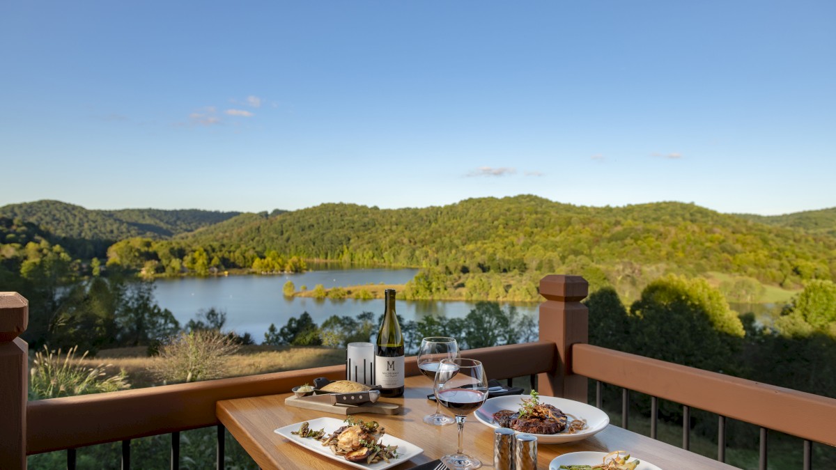 A table set for a meal overlooks a scenic landscape with a lake and lush green hills, featuring wine, two glasses, and plated food dishes.