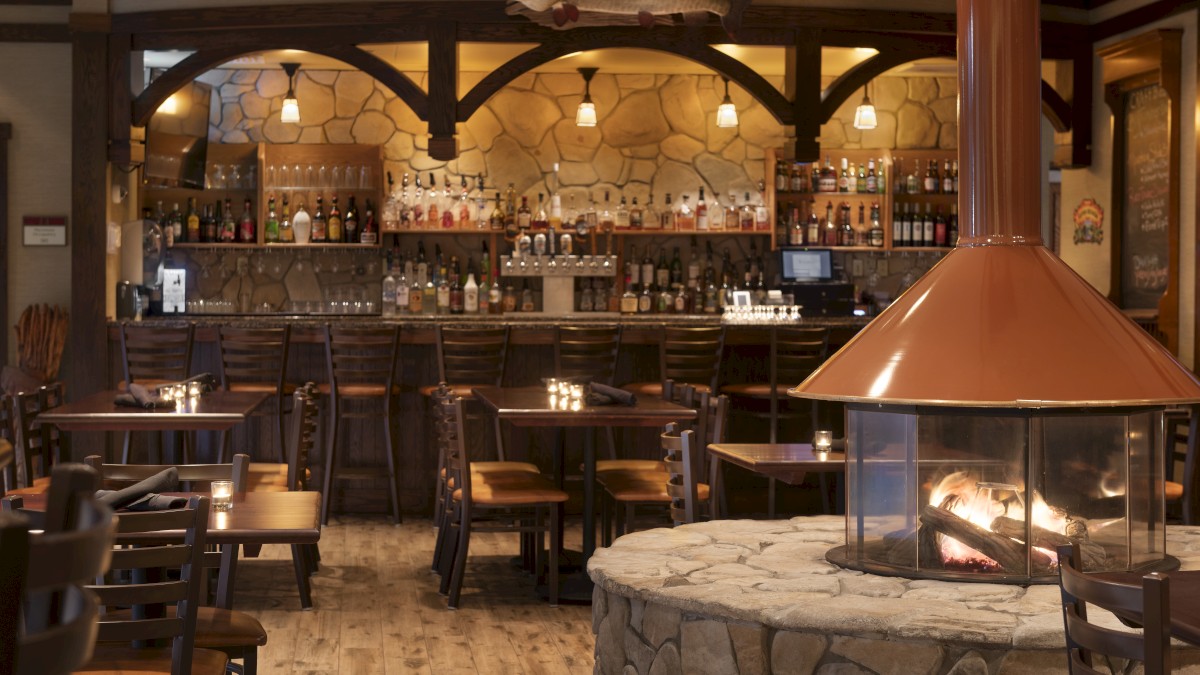 A cozy bar with a stone fireplace, wooden tables and chairs, and a well-stocked bar area. Warm lighting creates an inviting atmosphere.