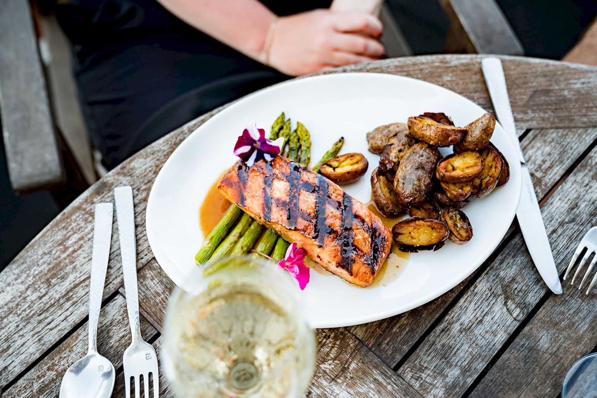 A plate of grilled salmon with asparagus and roasted potatoes on a wooden table, accompanied by a glass of white wine.