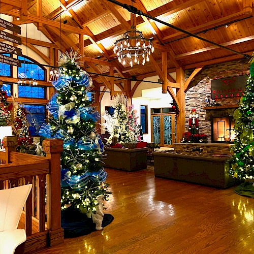 A cozy, festive indoor lounge with tall decorated Christmas trees, warm wooden beams, a stone fireplace, and comfortable seating in a lodge-style room.