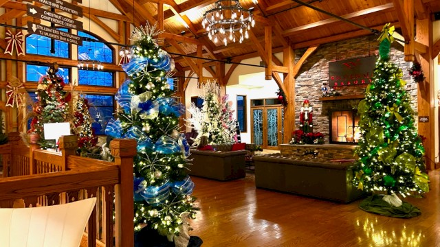 A cozy, festive indoor lounge with tall decorated Christmas trees, warm wooden beams, a stone fireplace, and comfortable seating in a lodge-style room.