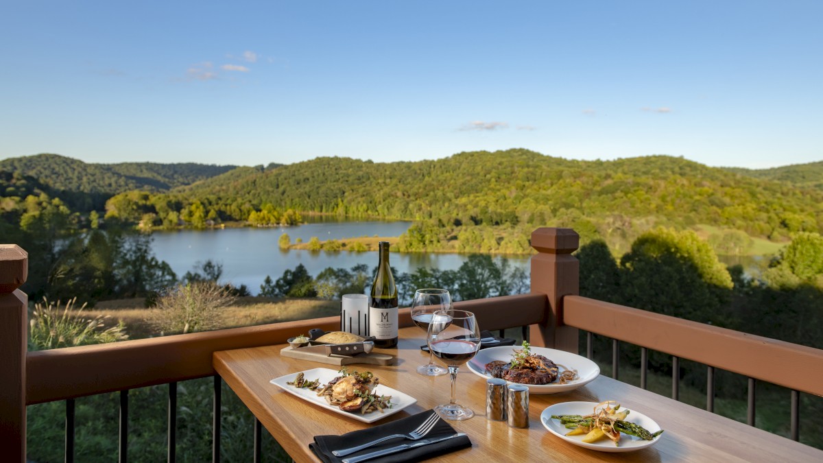 A dining table on a wooden balcony overlooks a serene lake and forested hills, set with plates of food, wine glasses, and a bottle of wine.