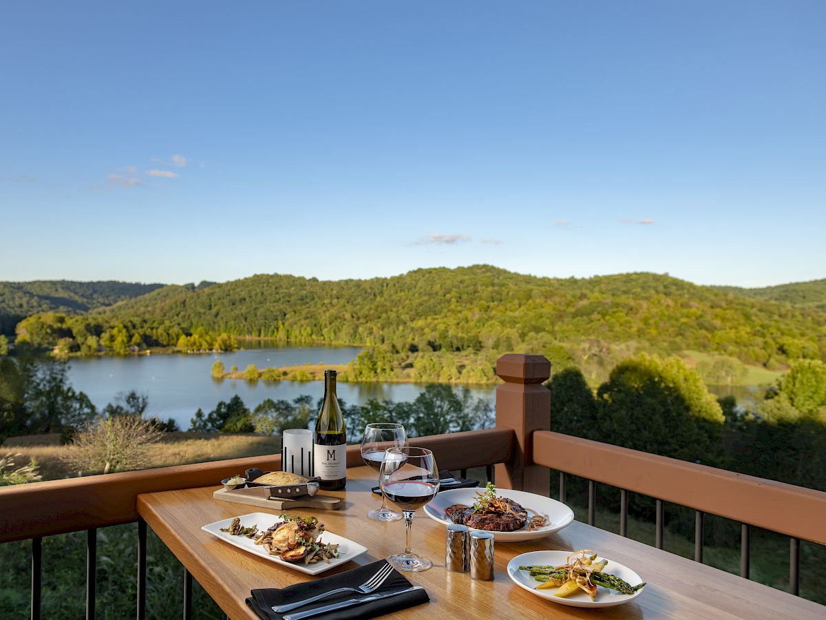 A dining table on a wooden balcony overlooks a serene lake and forested hills, set with plates of food, wine glasses, and a bottle of wine.