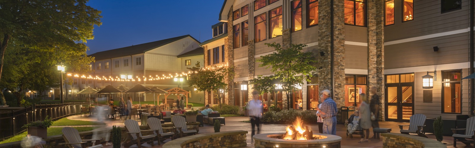The image shows a nighttime scene of a large, well-lit building with an outdoor seating area around a fire pit, with people socializing.