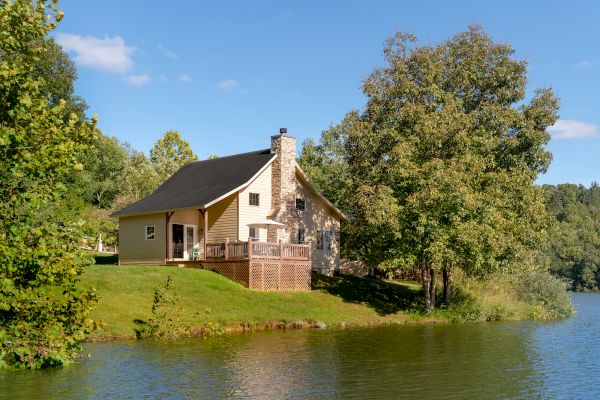 A house with a chimney and wooden deck is located by a calm lake, surrounded by trees and greenery, under a clear blue sky.