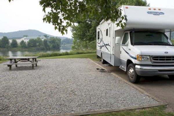 An RV is parked near a lakeside campground with a gravel parking area and a picnic table. Trees and hills are visible in the background.