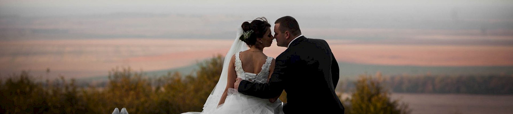 A bride and groom sit close on a hillside at sunset, sharing a kiss in a white wedding dress and black suit with a long veil and bouquet nearby.