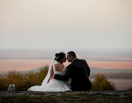 A bride and groom sit close on a hillside at sunset, sharing a kiss in a white wedding dress and black suit with a long veil and bouquet nearby.
