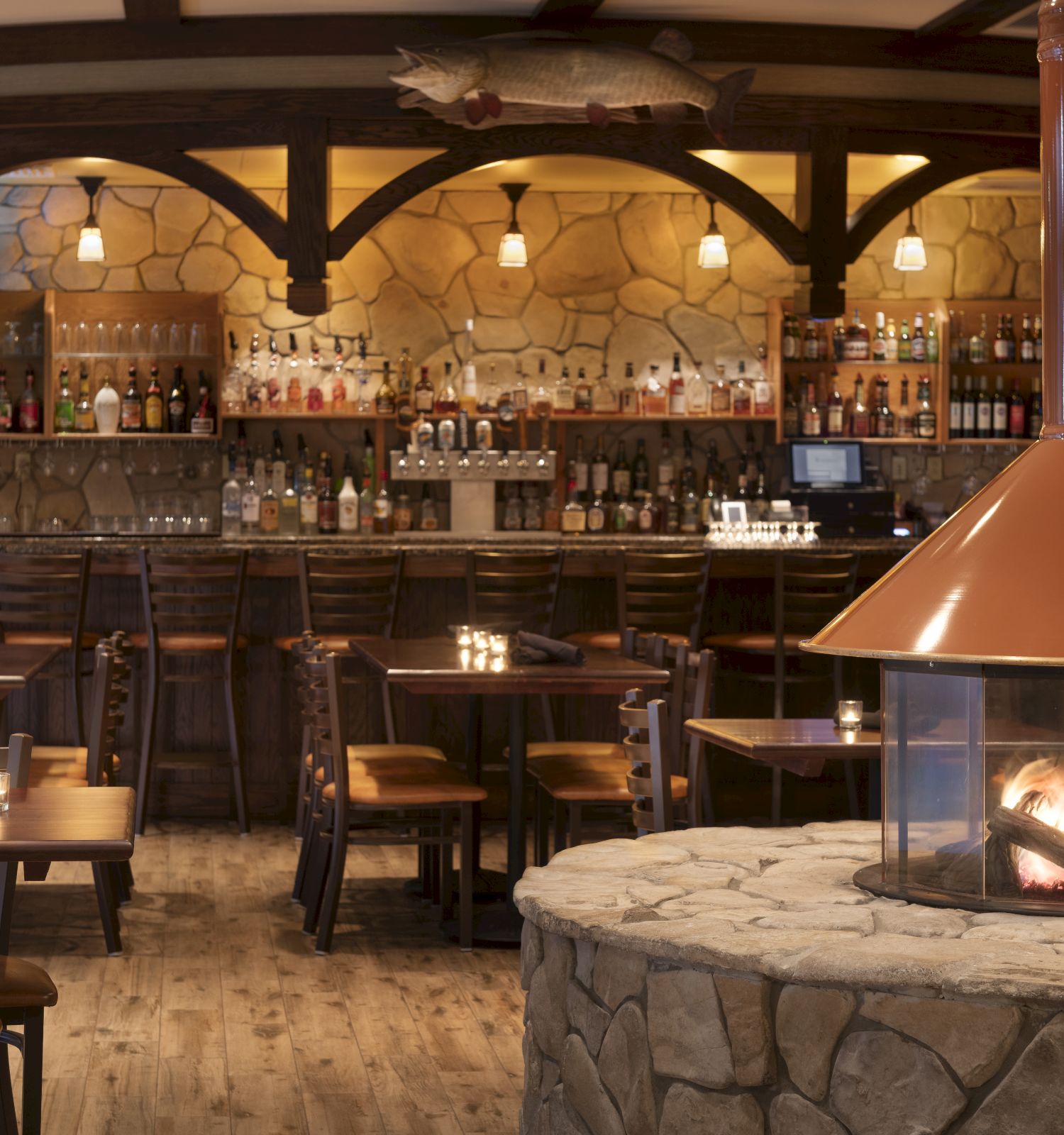 A cozy restaurant interior with wooden tables, chairs, a stone fireplace, and a fully stocked bar in the background, creating a warm, inviting atmosphere.