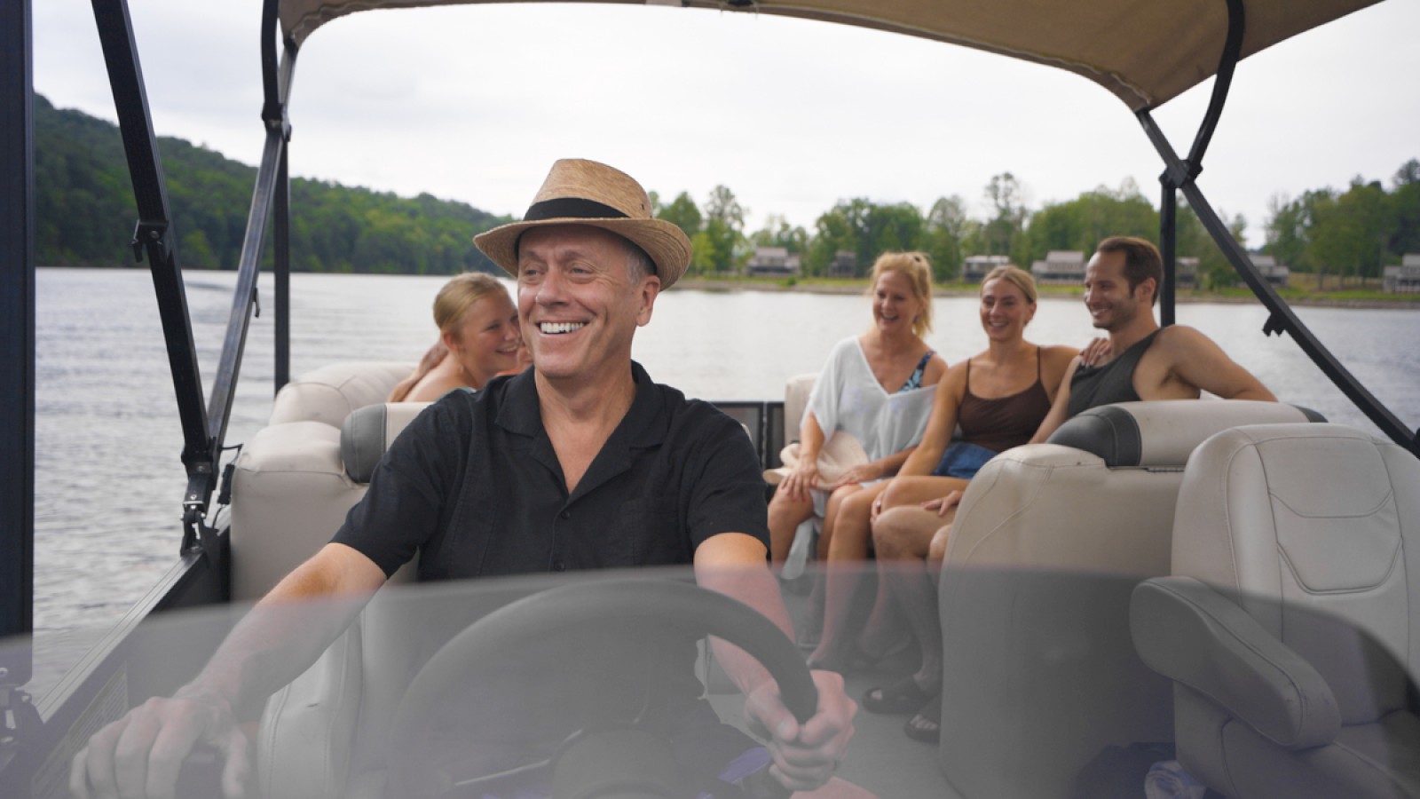 A man in a hat drives a boat with five smiling people enjoying a lake or river outing on a cloudy day.