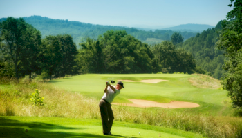 A golfer teeing off on a lush, hilly golf course with greens, sand traps, and a scenic forested backdrop.
