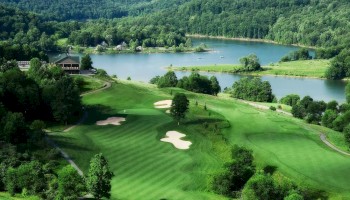 A lush, rolling golf course by a serene lake, with greens, sand bunkers, and surrounding trees and hills under a bright, clear sky.