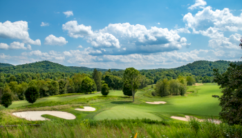 A lush golf course with well-manicured greens and sand bunkers, rolling hills, and a bright blue sky with fluffy clouds.