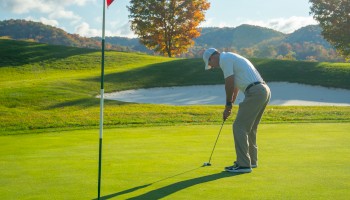 A golfer putting on a sunny green near a sand bunker and hills, with a flagstick and trees in the background.
