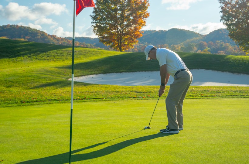 A golfer putting on a sunny green near a sand bunker and hills, with a flagstick and trees in the background.