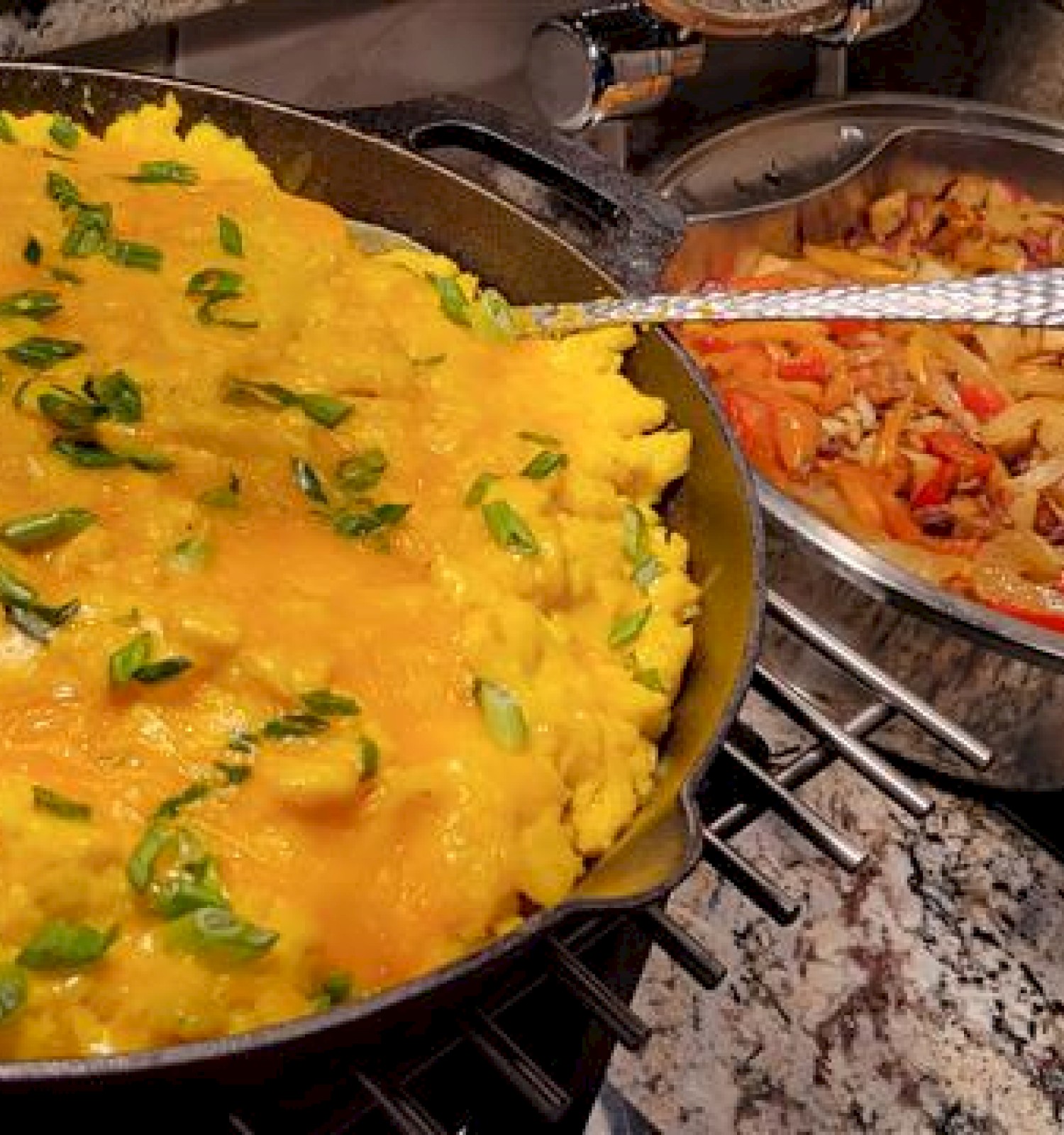 A large skillet of scrambled eggs with melted cheese and green onions next to a bowl of sautéed peppers and onions; both on a kitchen counter.