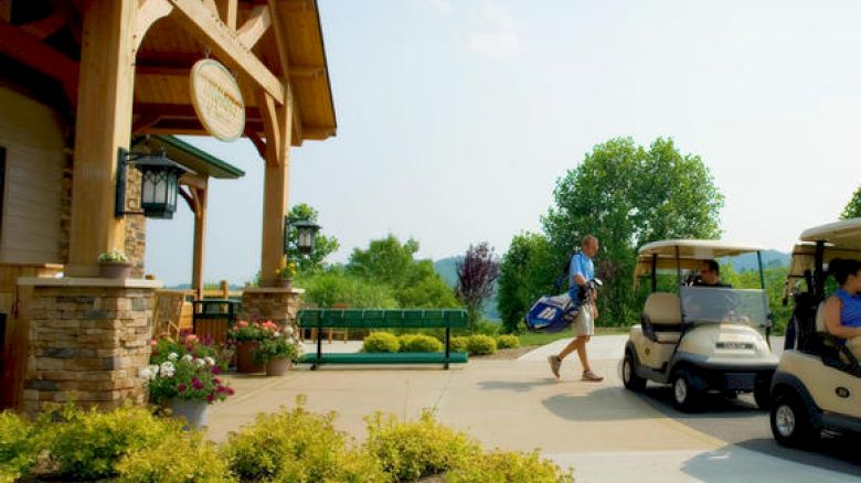 A person carrying golf clubs near a clubhouse, with two golf carts parked nearby and lush greenery surrounding the area.
