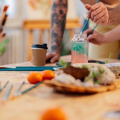 A messy art-and-snack table with brushes, a jar of water, a coffee cup, and a hand painting something while food sits nearby.